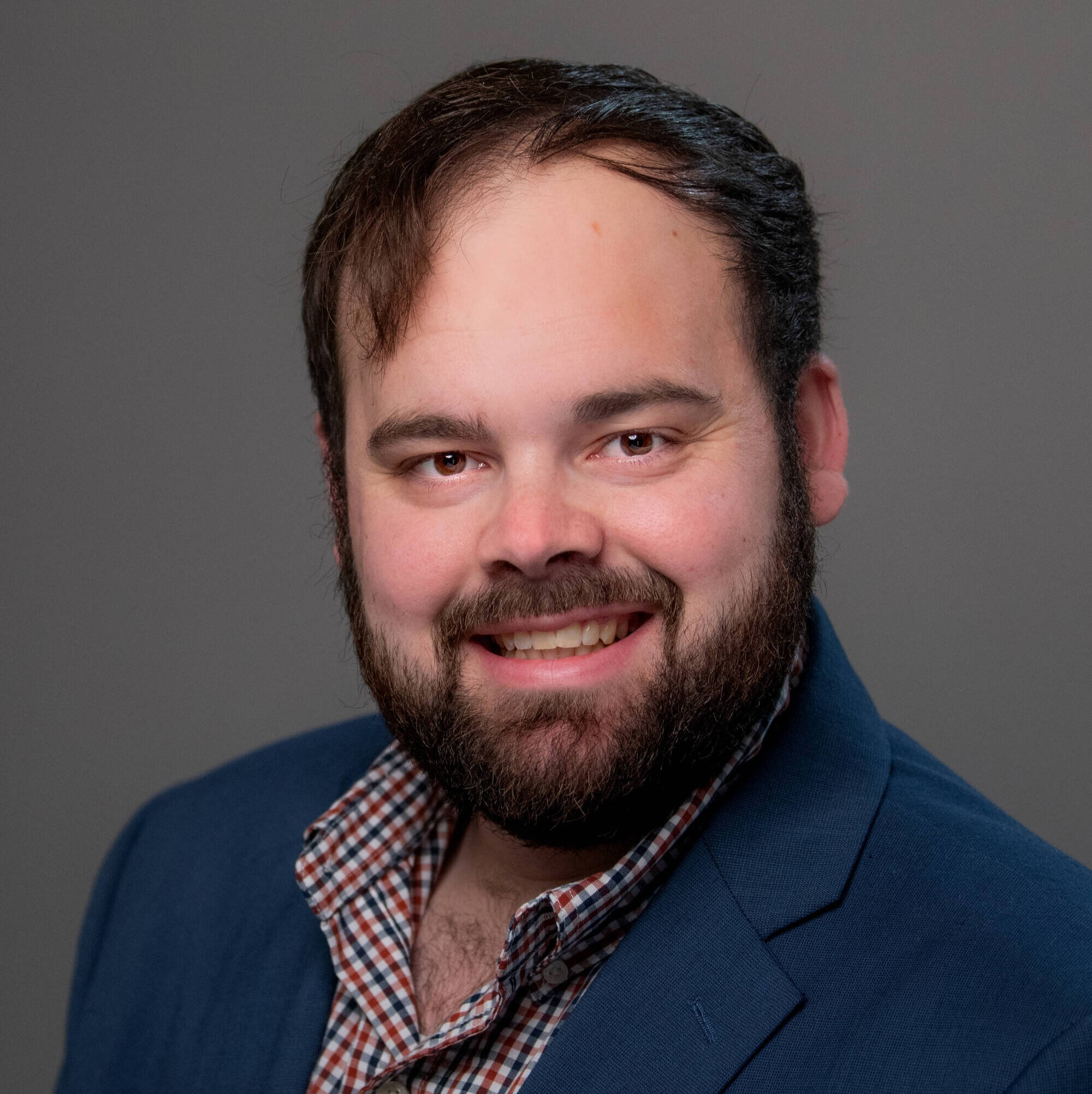 Brock Thane in a navy blazer with a plaid shirt on underneath smiling at the camera